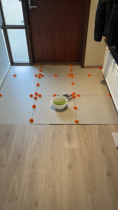 Kakelplattor placerade med nivåclips på golvet, en vit skål med grönt innehåll och en trasa ligger bland plattorna. En dörr i bakgrunden.