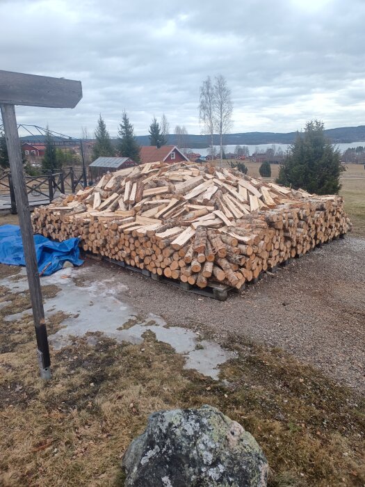 Stapel med ved på lastpallar i en trädgård, omgiven av landskap med träd och hus i bakgrunden.