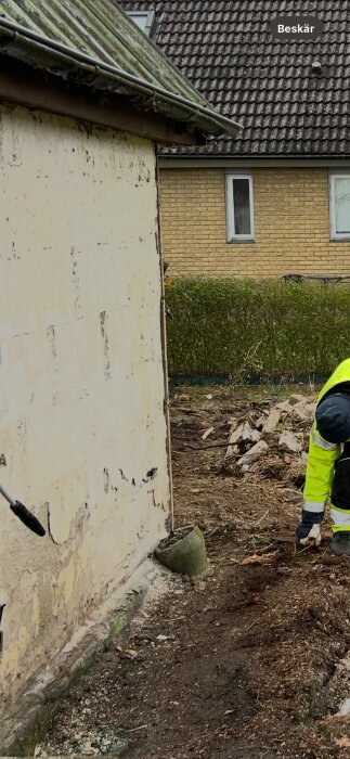 Skadad putsfasad med sprickor på husvägg. En person i arbetskläder arbetar med marken bredvid väggen.