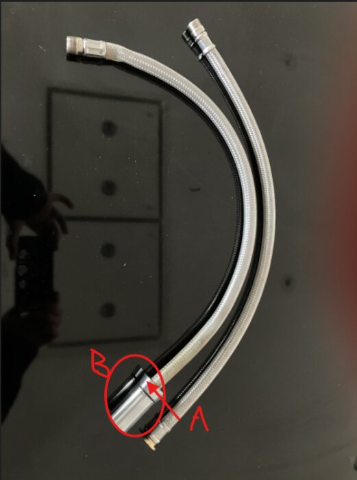 Två flätade slangar med en koppling markerad som "B" och en pil pekande mot kopplingen, mot svart bakgrund.