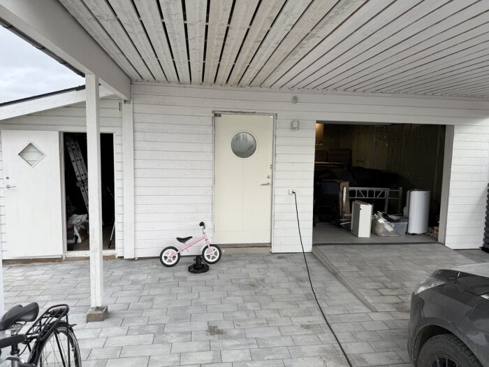 Vitt trähus med oisolerat garage, cykel och sparkcykel utanför. Garaget har öppen port och innehåller bland annat byggmaterial och verktyg.