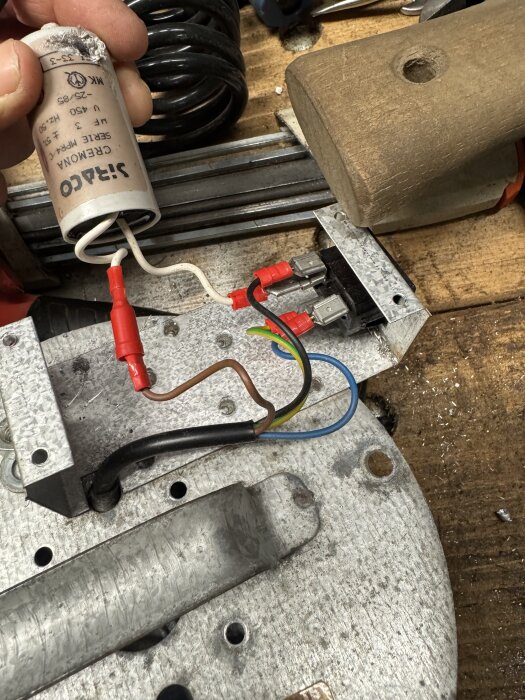 Elektrisk kondensator med flera anslutna kablar, liggandes på en arbetsbänk tillsammans med verktyg och metallkomponenter.