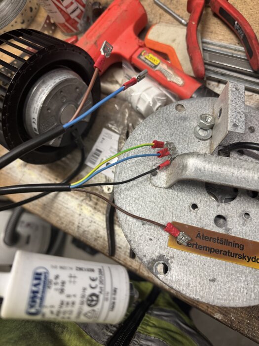 Bild av elektriska kablar och kontaktkopplingar på en fläktmotor bredvid en kondensator och verktyg på en arbetsbänk.