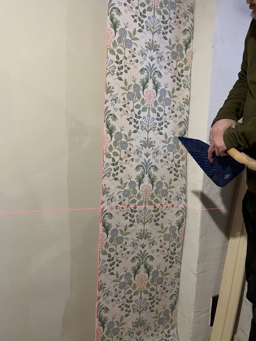 En person tapetserar en vägg med blommig tapet. En lasermätare används för att få tapeten rak.