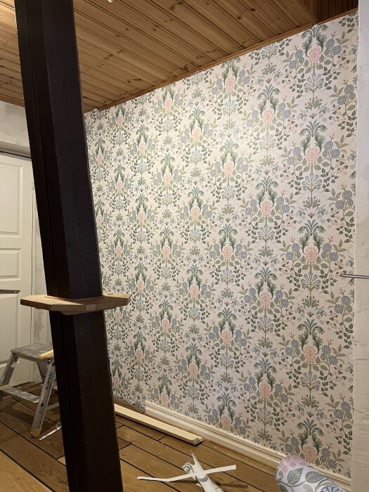 Vägg täckt med mönstrad tapet i blommigt motiv, träpanel i taket och träbjälke i rummet, en stege står vid sidan av väggen.