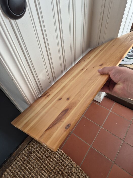 Person håller en träplanka mot en vit vägg för att planera ett hemmagjort skoställ med hyllplan. Plattans mått är 54 cm bredd och 18 cm djup.