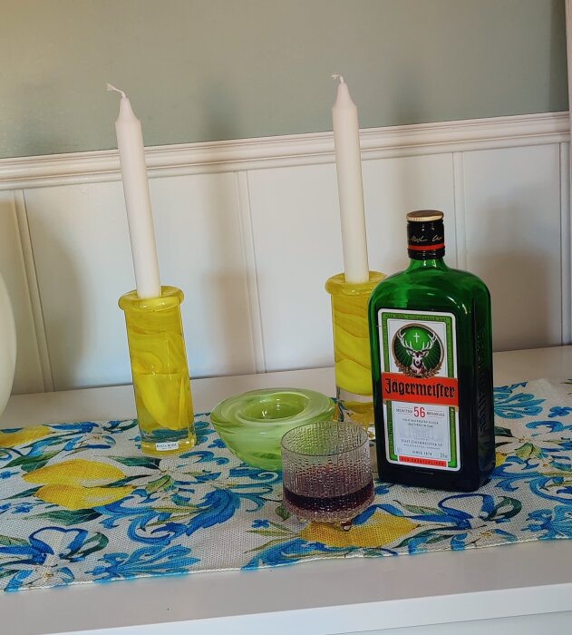 Flaska Jägermeister och glas bredvid ljusstakar på en mönstrad duk.