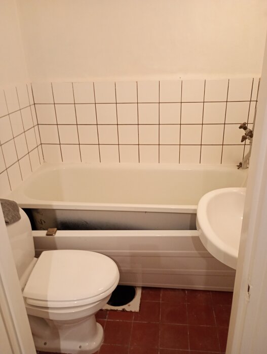 Äldre badrum med vitt kakel, inbyggt badkar, vit toalett och handfat. Rödbrunt golv, enkel inredning i behov av renovering.