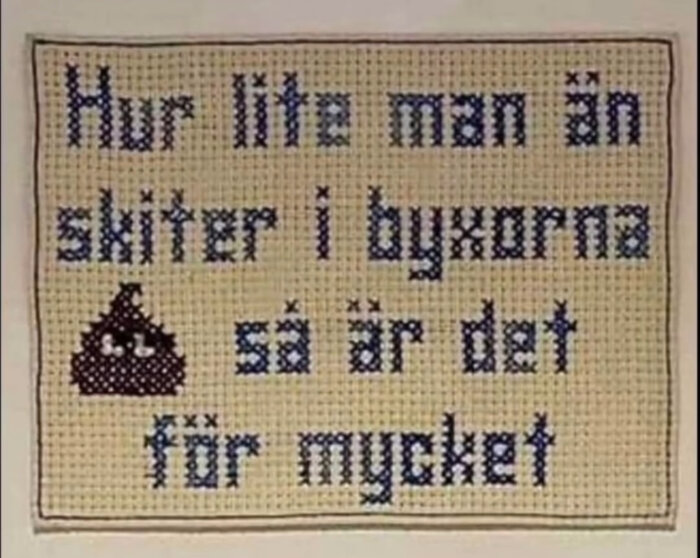 Väggbonad med korsstygnsbroderi innehåller humoristisk text och emoji-liknande motiv av bajshög med ögon.