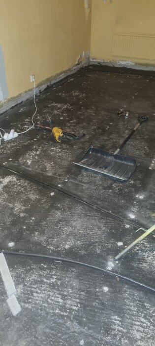 Svartmålat betonggolv med verktyg, kabel och snöskyffel i ett rum med avflagad färg på väggarna.