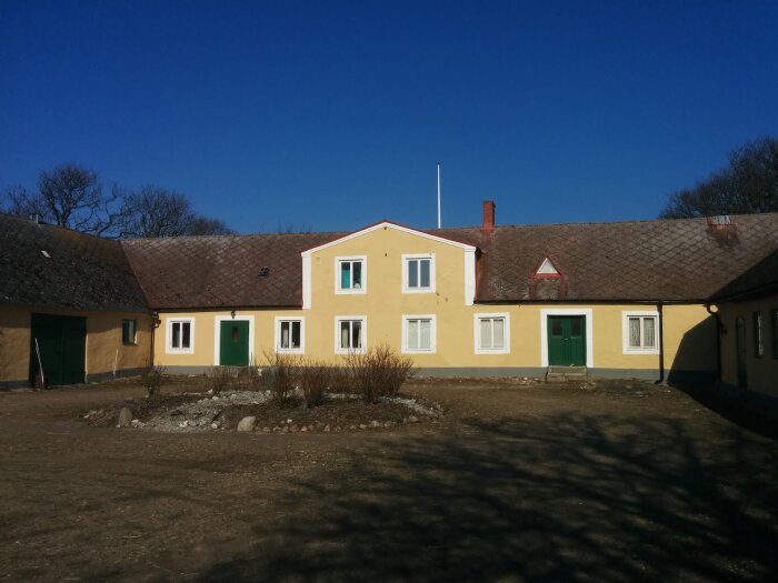 Gult boningshus i klassisk skånelänga-stil med tegeltak, gröna dörrar och vita fönster; omgivning av trädgård och klar himmel.