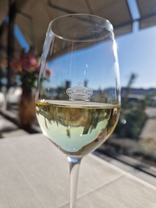 Glas med vitt vin med en krona på, står på ett bord utomhus under en klarblå himmel. Bakgrunden är suddig med blommor och grönska.