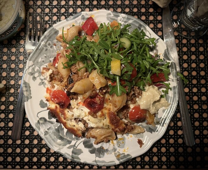 Köttfärsgratäng med pasta och mozzarella, toppad med sallad och ruccola, serverad på en tallrik med bestick på ett mönstrat underlägg.