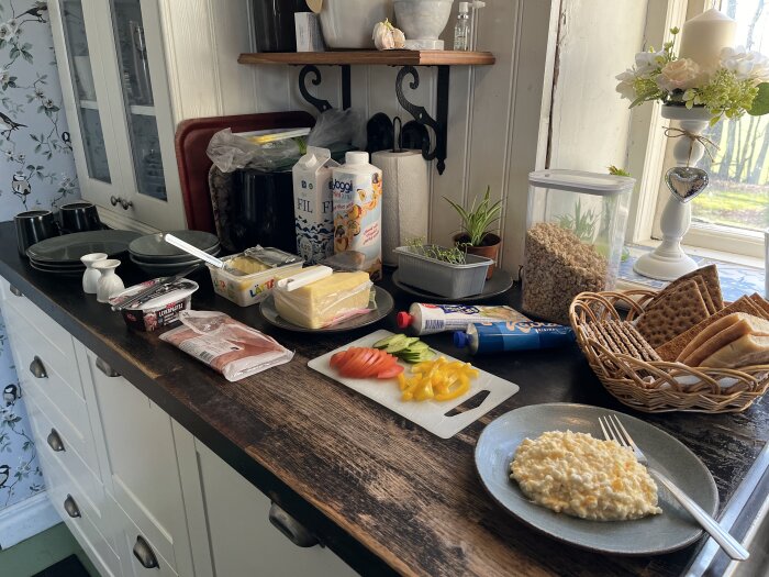 Frukostbord med tallrikar, pålägg, bröd och grönsaker. Ost, leverpastej, fil och knäckebröd finns på bänken i ett kök med blommig tapet.