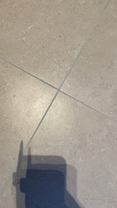 Ljusa kakelplattor på golvet med korsande fogar och en skugga i nedre delen av bilden.