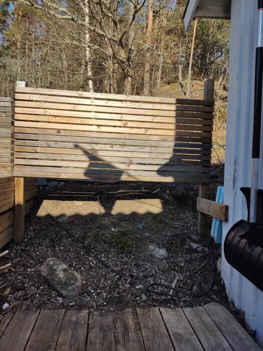 Trädäck mot sluttning med trästaket i bakgrunden samt mark med gräsfläckar och stenar. Del av husvägg syns till höger.