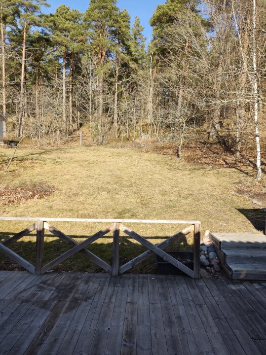 Altan med träräcke framför en gräsyta med sluttning och omgivande skog i bakgrunden under en klarblå himmel.