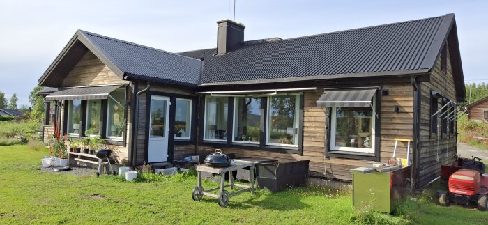 Trähus med järnvitriolbehandlad panel, omgärdat av gräs, med grill och trädgårdsdekoration framför huset.