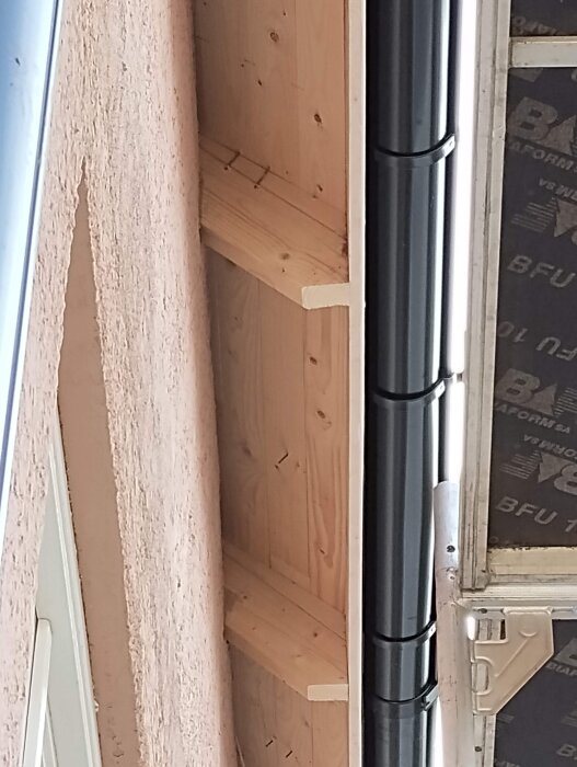 Obehandlad träfot ovanför ett fönster bredvid en svart hängränna, med oskyddad träkonstruktionsdetalj vid takfoten.