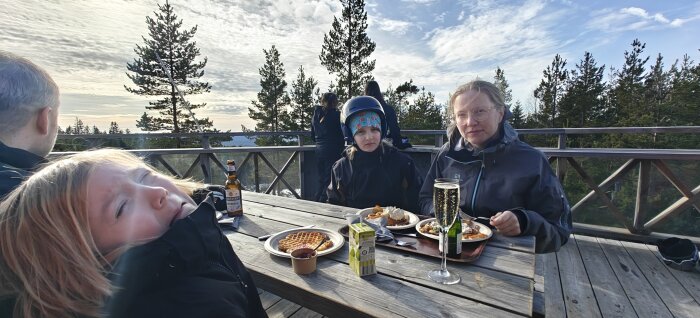 Personer sitter utomhus vid ett träbord och äter laxvåfflor, med skog i bakgrunden.