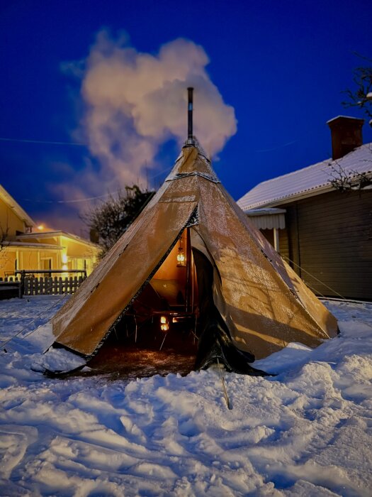 Tält i en snötäckt trädgård med rök från skorstenen och en upplyst interiör, kvällsbelysning från omkringliggande hus.