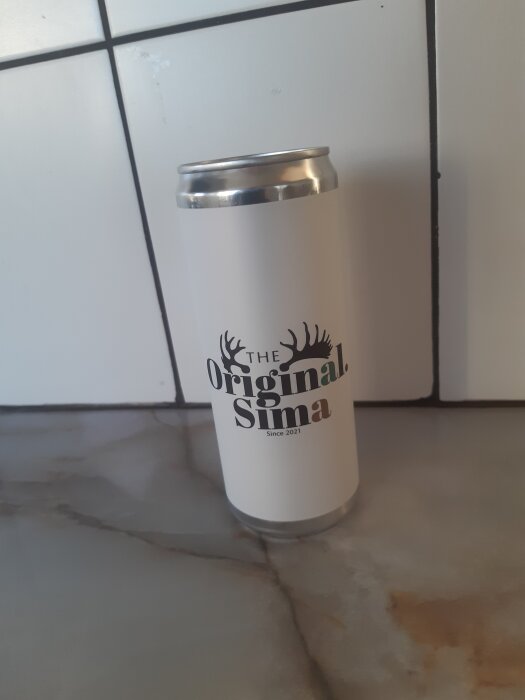 En vit dryckesburk märkt "The Original Sima Since 2021" står på ett bord med marmormönster och en kaklad vägg i bakgrunden.