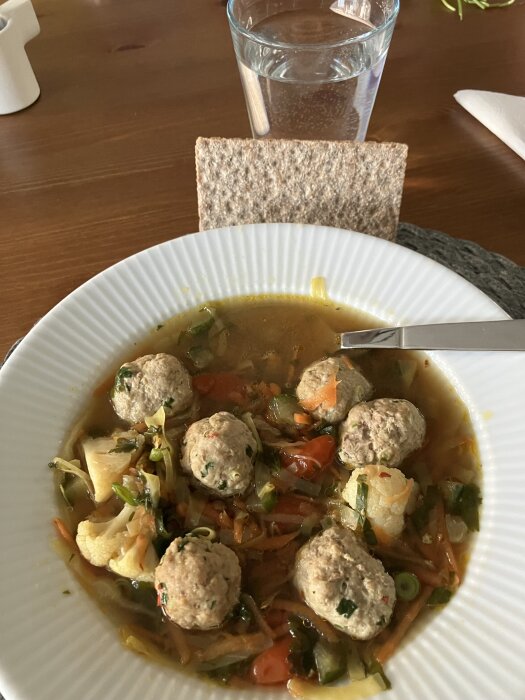 Grönsakssoppa med asiatiska frikadeller serverad i en vit tallrik, bredvid ett glas vatten och en skiva knäckebröd.