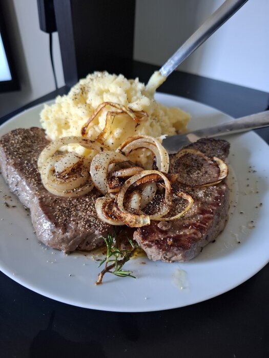 Rätt lyxig fredagsmiddag med ryggbiff, stekt lök och potatismos, serverad på en tallrik med kniv och gaffel bredvid.