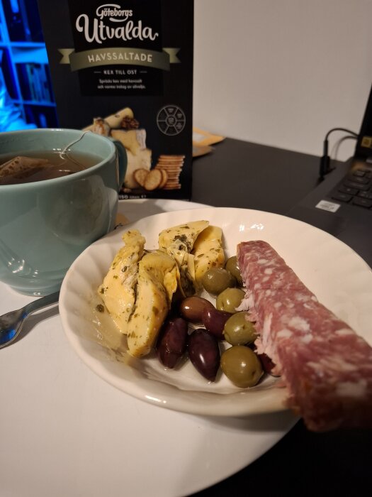 En tallrik med ost, oliver och en bit salami bredvid en kopp te och kexförpackning.