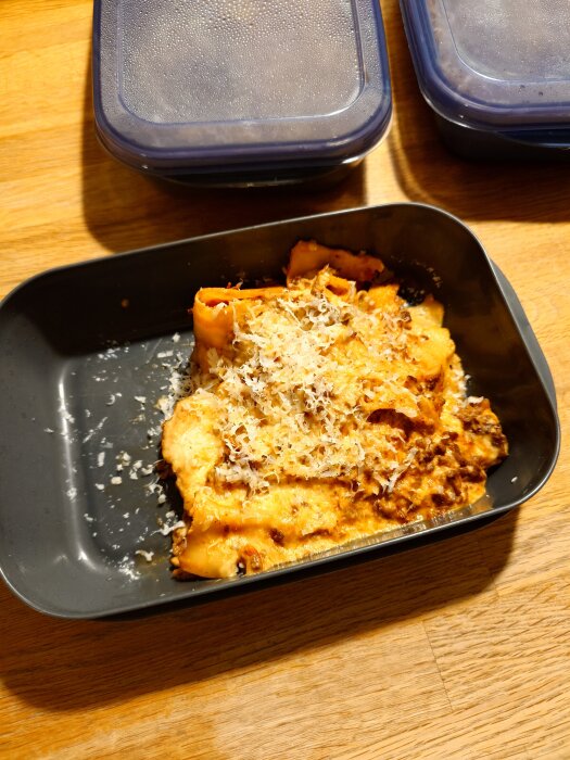 Lasagne i svart ugnsform med riven ost på ett träbord, tillsammans med matlådor med lock.