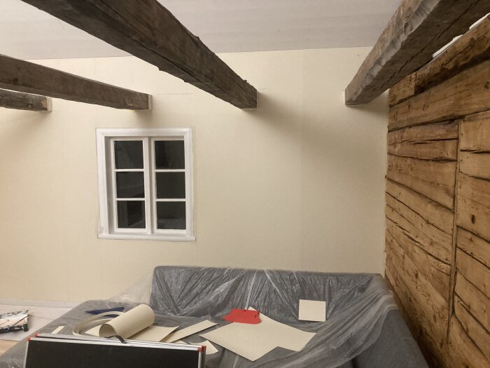 Ett rum under renovering med nytapetserade väggar, exponerade träbjälkar i taket och spånskivor samt plast övertäckt möbel.