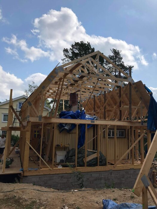 Byggarbetsplats med hus under konstruktion, visar trästomme och oslagna takstolar mot en blå himmel och träd i bakgrunden.