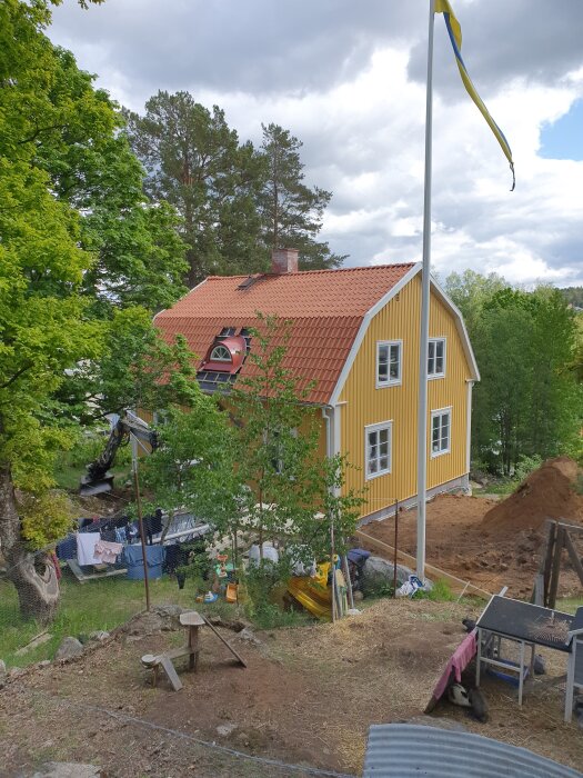 Gult trähus med rött tegeltak omgiven av träd, svensk flagga vajande i vinden, grävmaskin och byggmaterial synligt i trädgården.