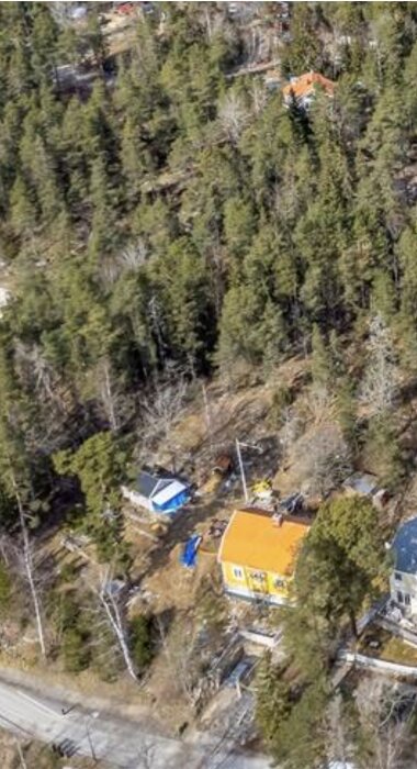 Flygfoto över ett gulmålat hus med orange tak omgivet av tät skog och en väg i förgrunden.