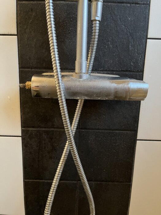 Silverfärgad duschblandare framför kakelvägg, med anslutna flexibla slangar.