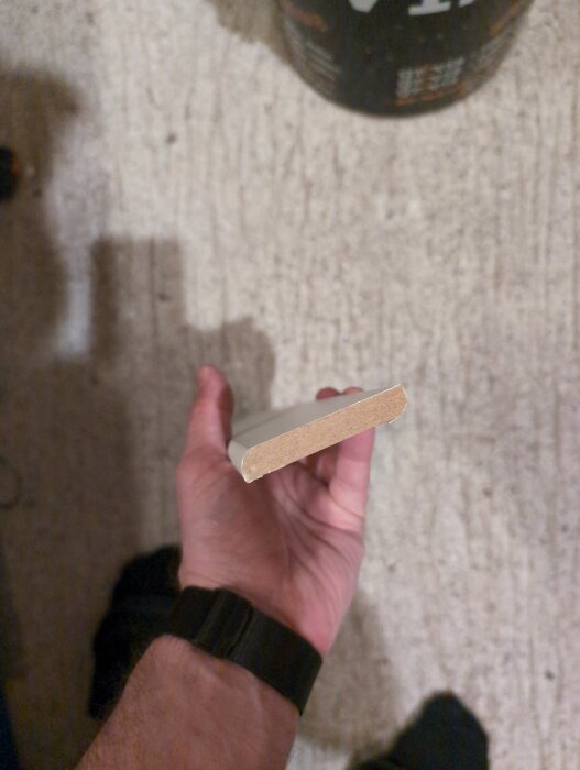 Närbild av en hand som håller i en vit laminat golvlist, 60 mm hög och 5 mm tjock, mot en ljus bakgrund.
