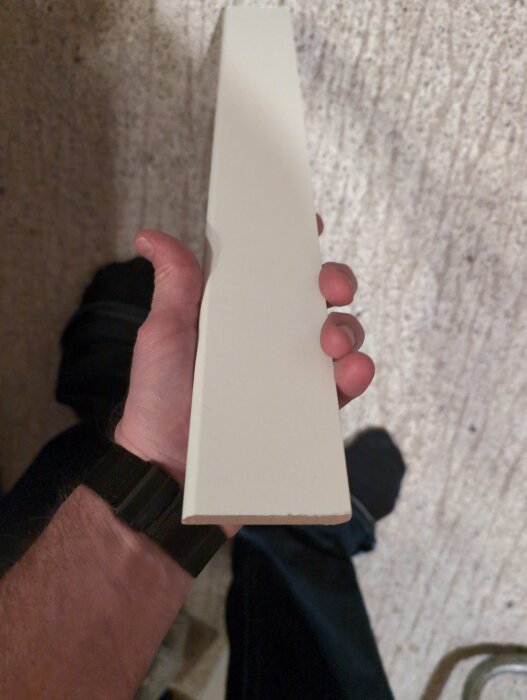En person håller en vit laminatgolvlist i handen, 60 mm hög och 5 mm tjock, mot en ljus bakgrund.
