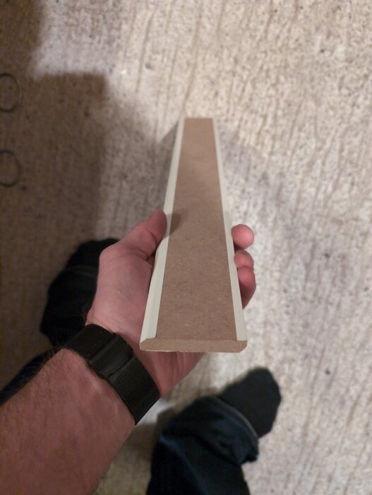 En hand håller en brun, enkel laminatgolvlist med vit kant, mot en beige bakgrund. Listen är 60 mm hög och 5 mm tjock.