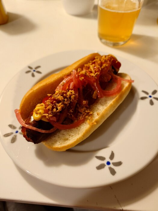 Grillkorv i bröd med senap, ketchup, picklad rödlök och rostad lök på en tallrik, bredvid ett glas öl.
