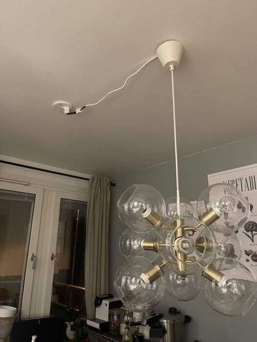 Pendellampa med nio E14 socklar hänger från taket, ansluten med sladdar till ett lamputtag. Bakgrund med fönster och gardiner syns.