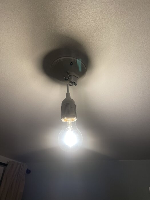 En tänd glödlampa i ett enkelt takfäste, med synlig sladd och vit takplatta.
