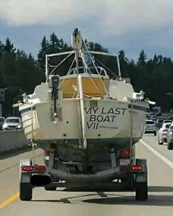 Båt med texten "My Last Boat VII" transporteras på en släpvagn längs en väg, omgiven av träd och trafik.