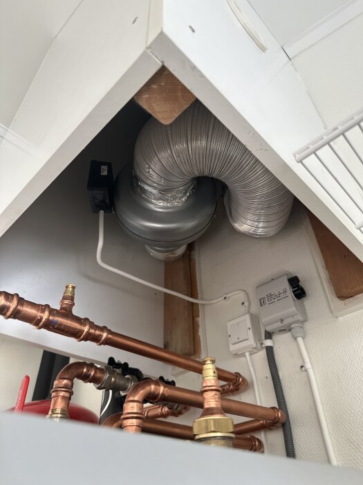 Ventilationsinstallation med en kanalfläkt i ett rum, silverrör anslutet till fläkten i hörnet. Kopparrör och elektriska kablar visas också.