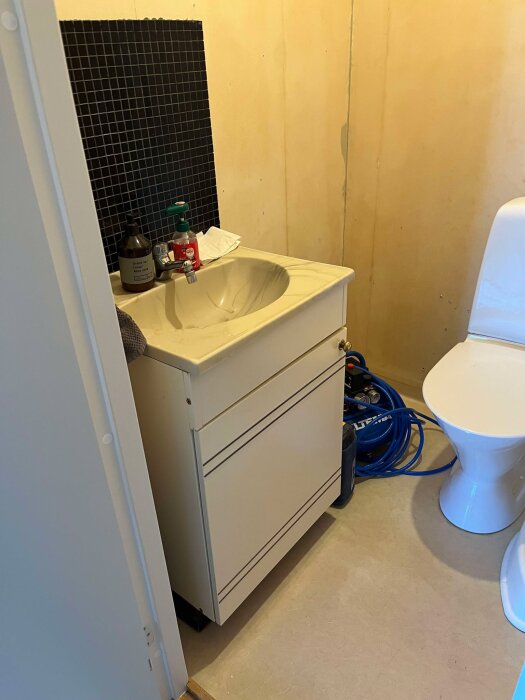 Litet badrum med beige väggar, vit tvättställs möbel, svart kakelpanel bakom och toalett. Golvet har en våtrumsmatta.