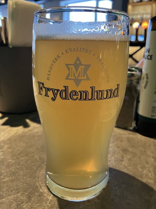 Ett glas öl med "Frydenlund" tryckt på, stående på ett bord i ett belyst rum.