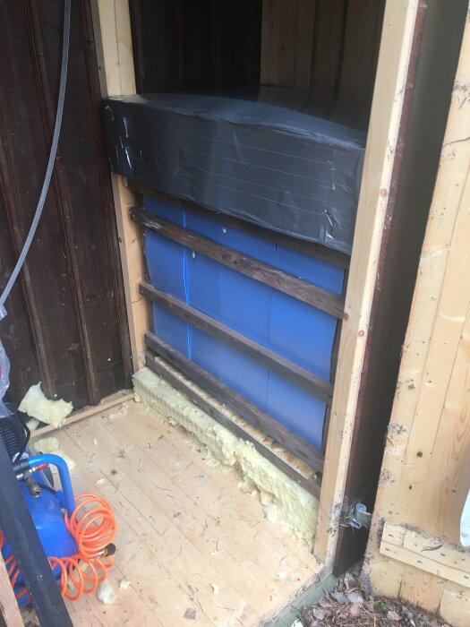 Isolerad ram i ett uthus med blå isolering och underlagsskivor, silvertejp runt ett lock, synlig orange sladd på golvet.