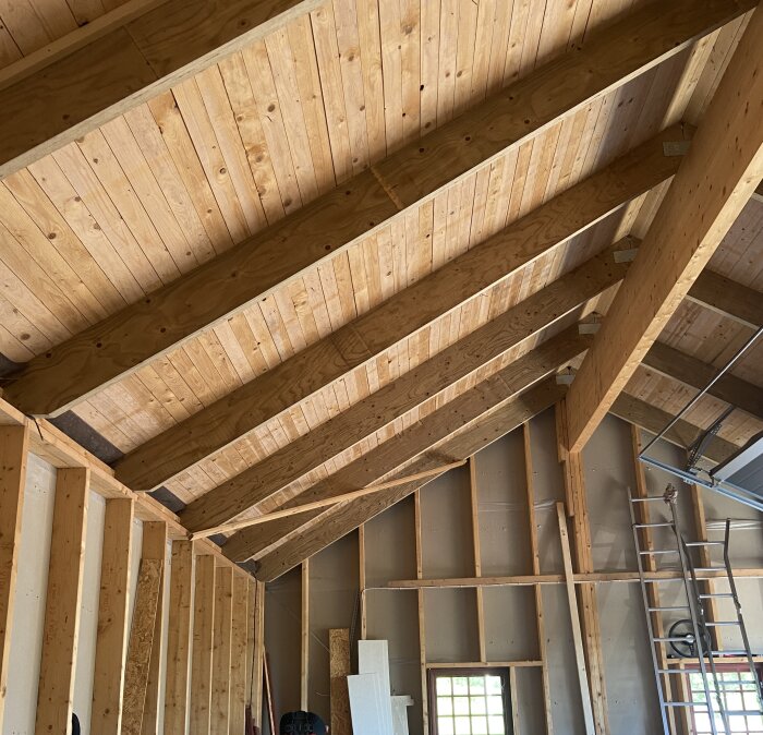 Garagetak med träpanel och öppet till nock, synliga takbjälkar och halvfärdiga väggar, stege lutad mot väggen.