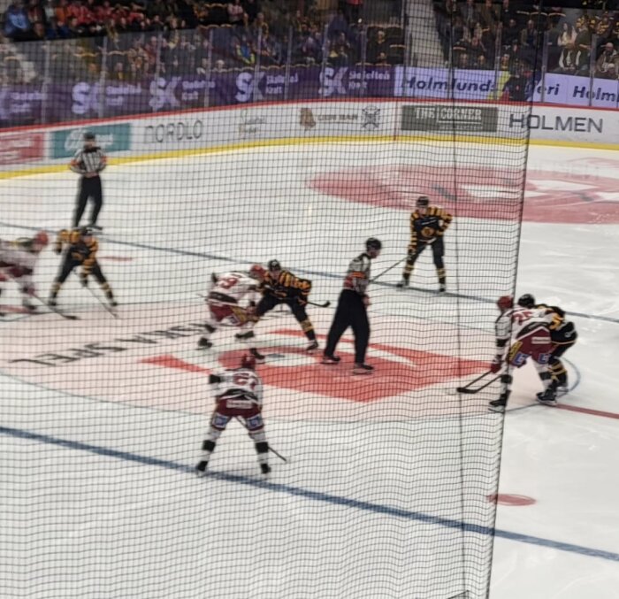 Ishockeymatch pågår med spelare i röda och svarta tröjor som gör sig redo för nedsläpp på isen, domaren står mellan två spelare.