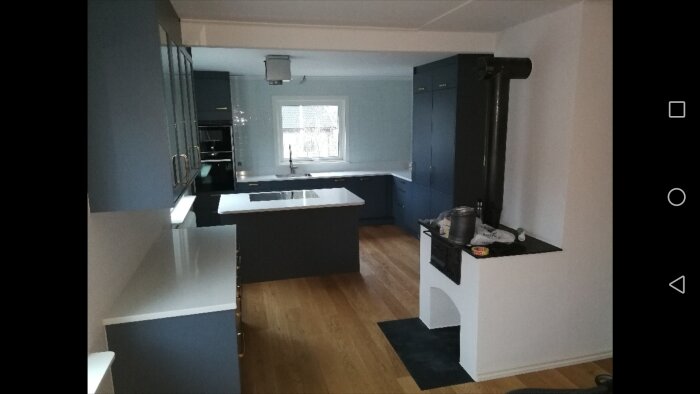 Modernt Marbodal-kök med blåa skåp, vit bänkskiva, fönster och en kamin. Trägolv och centralt placerad köksö.