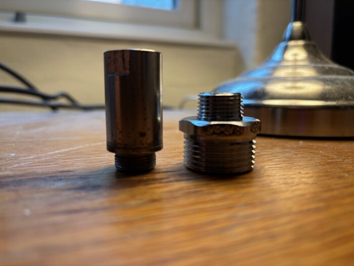 Två metallnipplar på ett bord; den vänstra med smalare gängor, den högra med grövre gängor. Bakgrund med lampa och fönster.